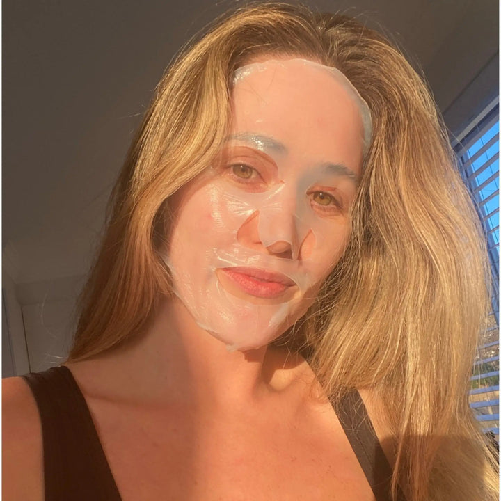 Woman with a bio-cellulose sheet mask on her face, sunlit room with blinds casting shadows, enjoying a self-care moment in Australia. Shop at VAMS beauty Biodegradable Bio Cellulose masks.