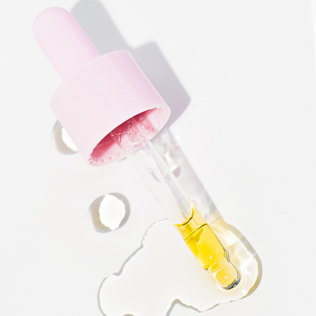 Close-up of a pink dropper from Oatbody dispensing yellow face oil, creating a shiny outline on a bright white background, highlighting the product's pure and hydrating oil texture.