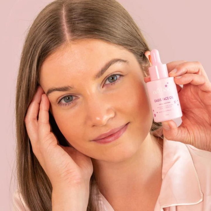 Young woman with fair skin and light brown hair holding a pink bottle of Oatbody Oasis Face Oil, demonstrating the product's use on her flawless skin, set against a soft pink background to highlight its suitability for all skin types.