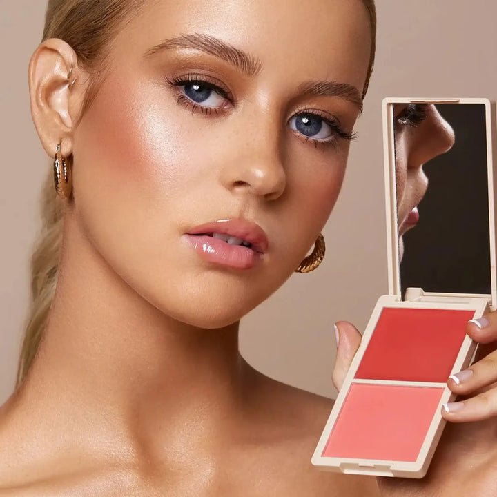 Model showcasing a radiant makeup look while holding the Make Me Blush Cream & Powder Duo in the shade 'Flirty.' The compact features a vibrant cream blush and matching powder, perfect for achieving a natural, glowing complexion. Ideal for creating versatile and long-lasting cheek colour.