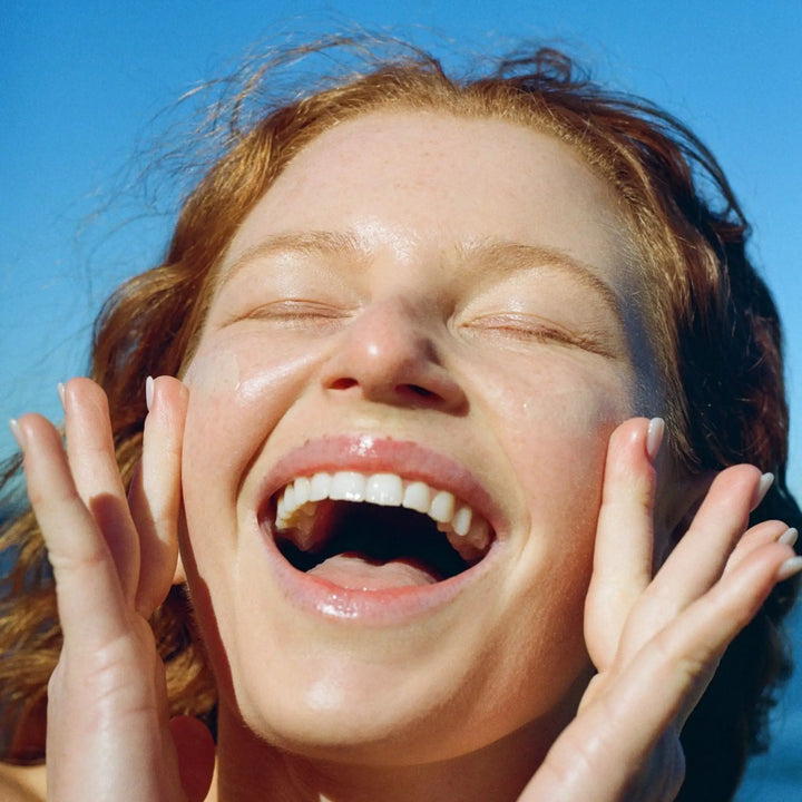 woman with glowing, radiant skin, smiling joyfully under a clear blue sky, highlighting the effects of Mother SPF mineral sunscreen for oily and acne-prone skin.