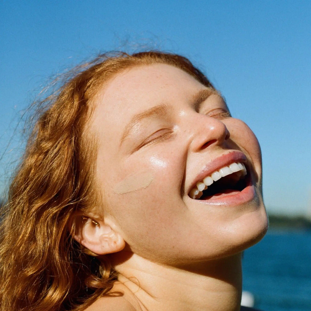 Woman with fair skin and red hair smiling under the sun, showing a swipe of Mother SPF mineral sunscreen for oily and acne-prone skin on her cheek, enjoying radiant skin protection with a natural, lightweight finish.