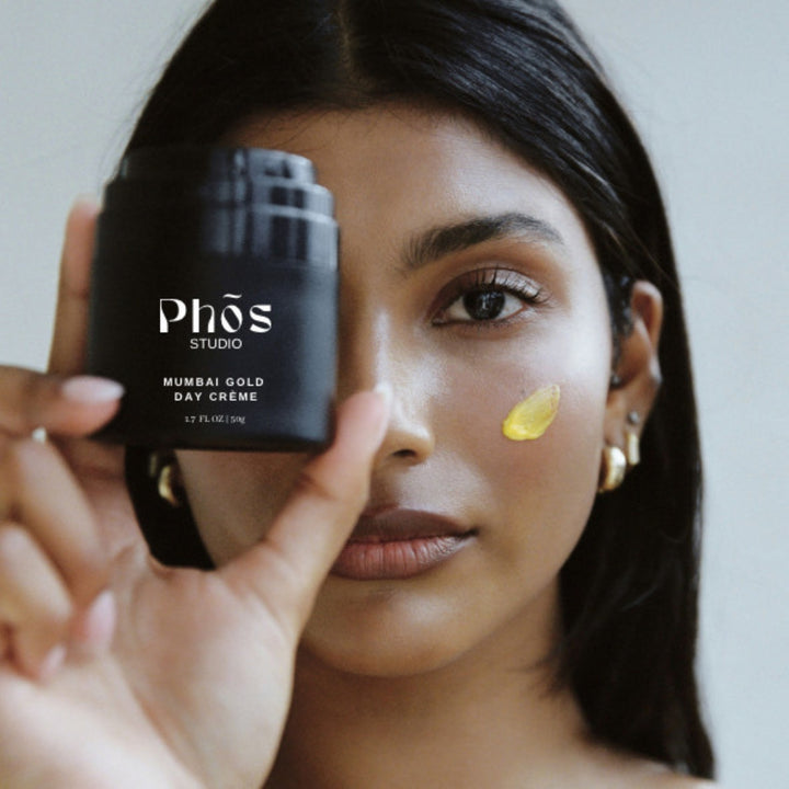 Portrait of a South Asian woman holding Phōs Studio Mumbai Gold Day Crème with a dab of yellow cream on her cheek, showcasing the product's texture and application method.