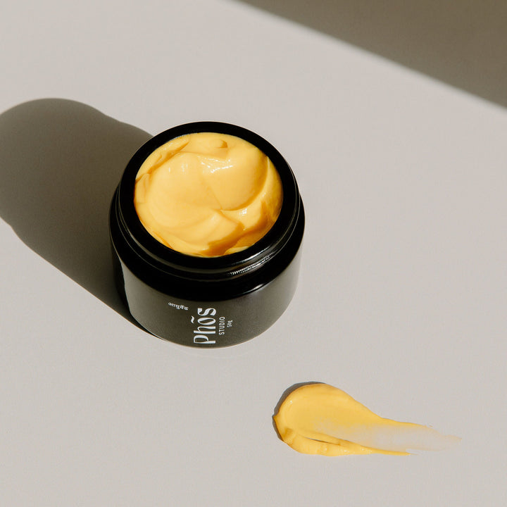 Open container of Phōs Studio Mumbai Gold Day Crème displaying its rich yellow texture, set against a contrasting shadow on a bright white surface, highlighting the product's luxurious consistency.