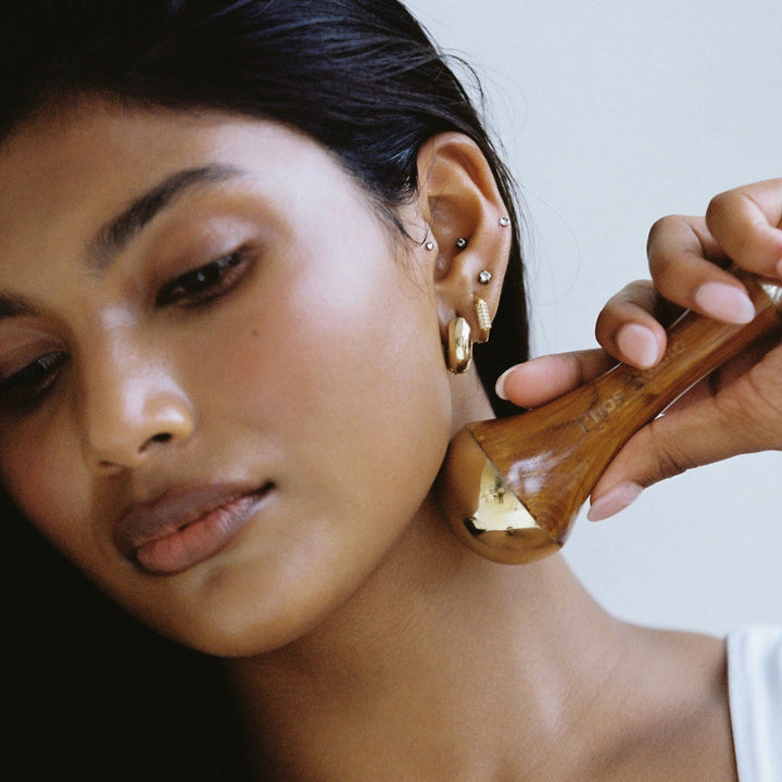 A close up of the Kansa Wand being held by a model, a traditional Ayurvedic facial massage tool made from a blend of copper and tin, with a wooden handle, used for detoxifying and soothing the skin.