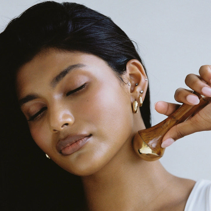 A close up of the Kansa Wand being held by a model, a traditional Ayurvedic facial massage tool made from a blend of copper and tin, with a wooden handle, used for detoxifying and soothing the skin.