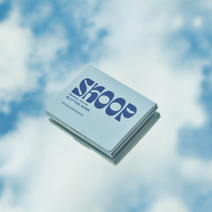 Skoop Babe Blotter Extra Drip Charcoal Blotting Paper in light blue packaging, reflected against a mirrored surface with a serene sky backdrop. Designed with 100 charcoal-infused sheets, these blotting papers provide a portable and stylish solution for oil control and skin freshness.