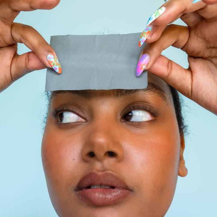 Model using Skoop Babe Blotter Extra Drip Charcoal Blotting Paper on her forehead, demonstrating its oil-absorbing effectiveness. The close-up highlights her glowing skin and colourful nail art, set against a soft blue background, showcasing a fresh and vibrant skincare routine.