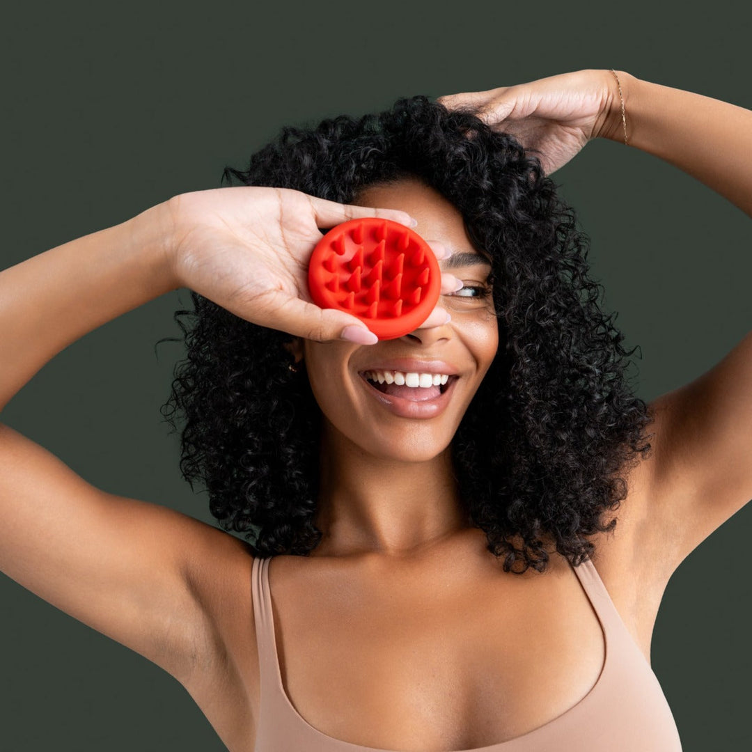 Smiling woman with curly hair holding OVEV Wellness red scalp scrubber, promoting hair growth and scalp care for all hair types