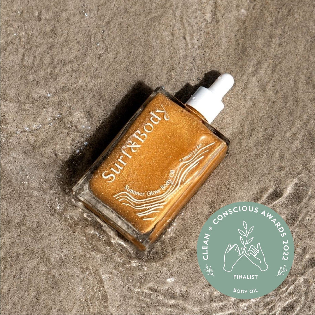 Summer Glow Body Oil bottle resting on wet sand, awarded as a finalist in the Clean + Conscious Awards 2022 for the body oil category, highlighting its natural, shimmering formula designed to enhance skin with a radiant glow.