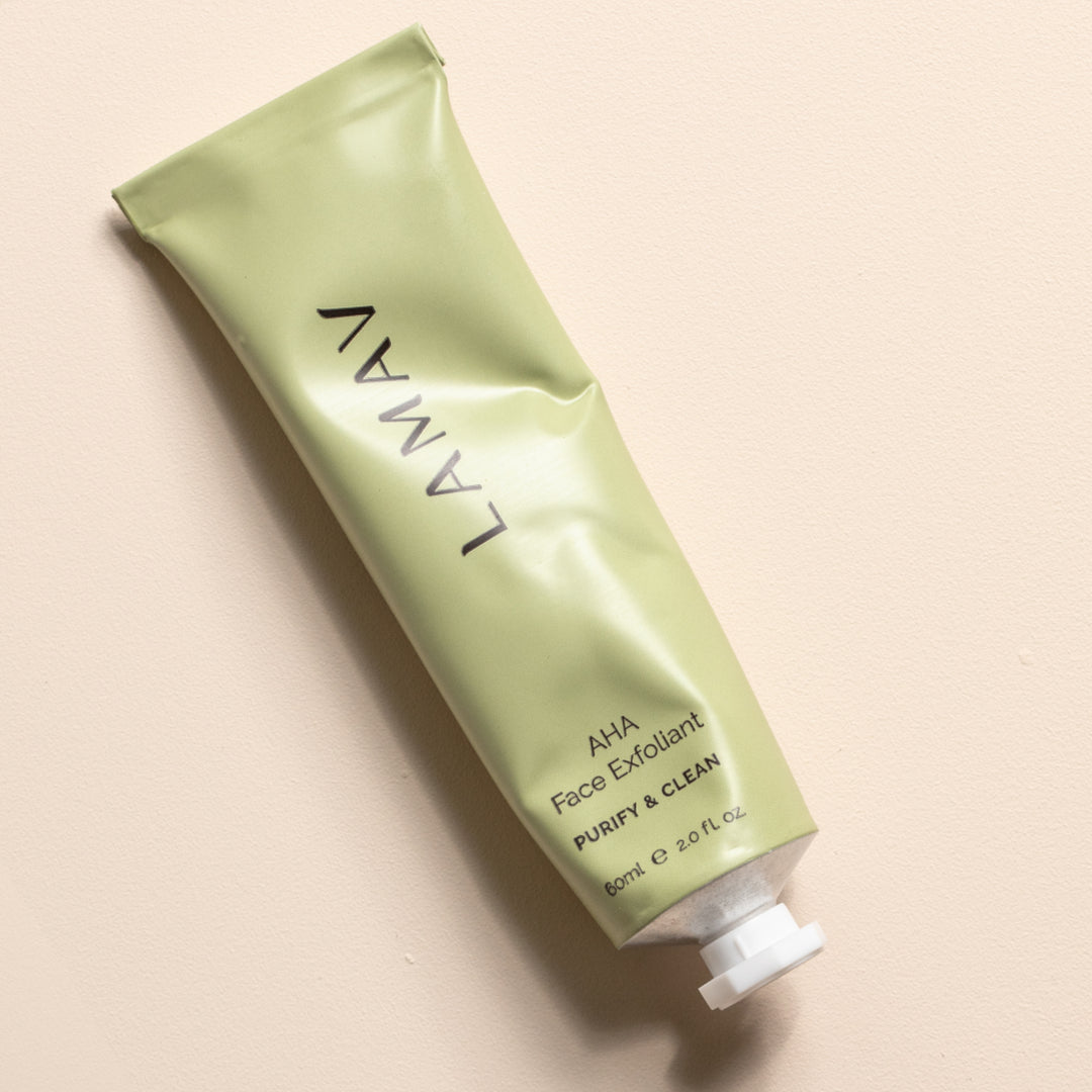 A slanted tube of LAMAV AHA Face Exfoliant lies against a light background with a smear of the product next to it, emphasizing its creamy texture.