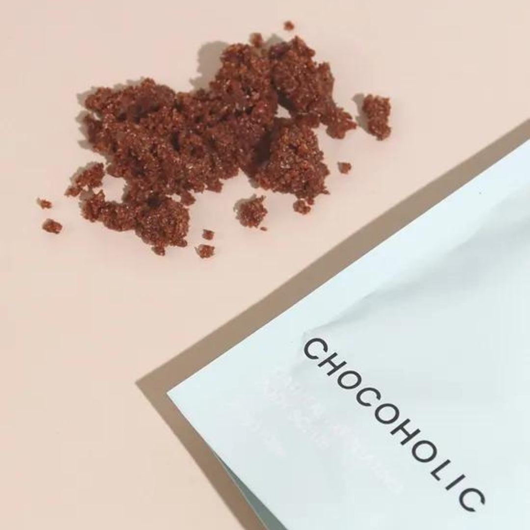 A bag of Oatbody Skincare's 'Chocoholic' body scrub with a playful warning 'CAUTION… I BREAK FOR CHOCOLATE,' against a soft pink background. The product is a gentle body scrub, this product help with bumpy, rough, and dry skin. 