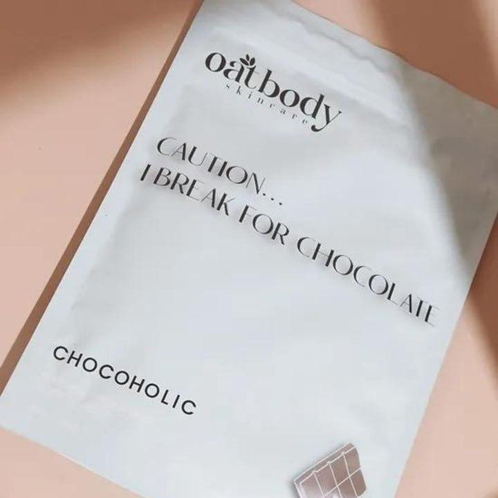 A bag of Oatbody Skincare's 'Chocoholic' body scrub with a playful warning 'CAUTION… I BREAK FOR CHOCOLATE,' against a soft pink background. The product is a gentle body scrub, this product help with bumpy, rough, and dry skin. 