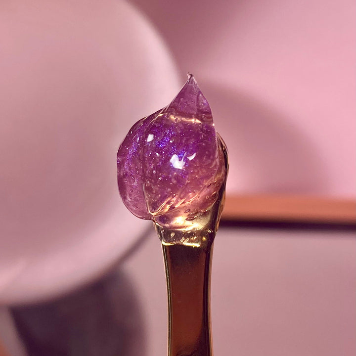 Close-up of a golden skincare tool with a drop of shimmering purple Muse by Gaia jelly exfoliant against a soft pink background, emphasizing the product's unique texture and visual appeal.
