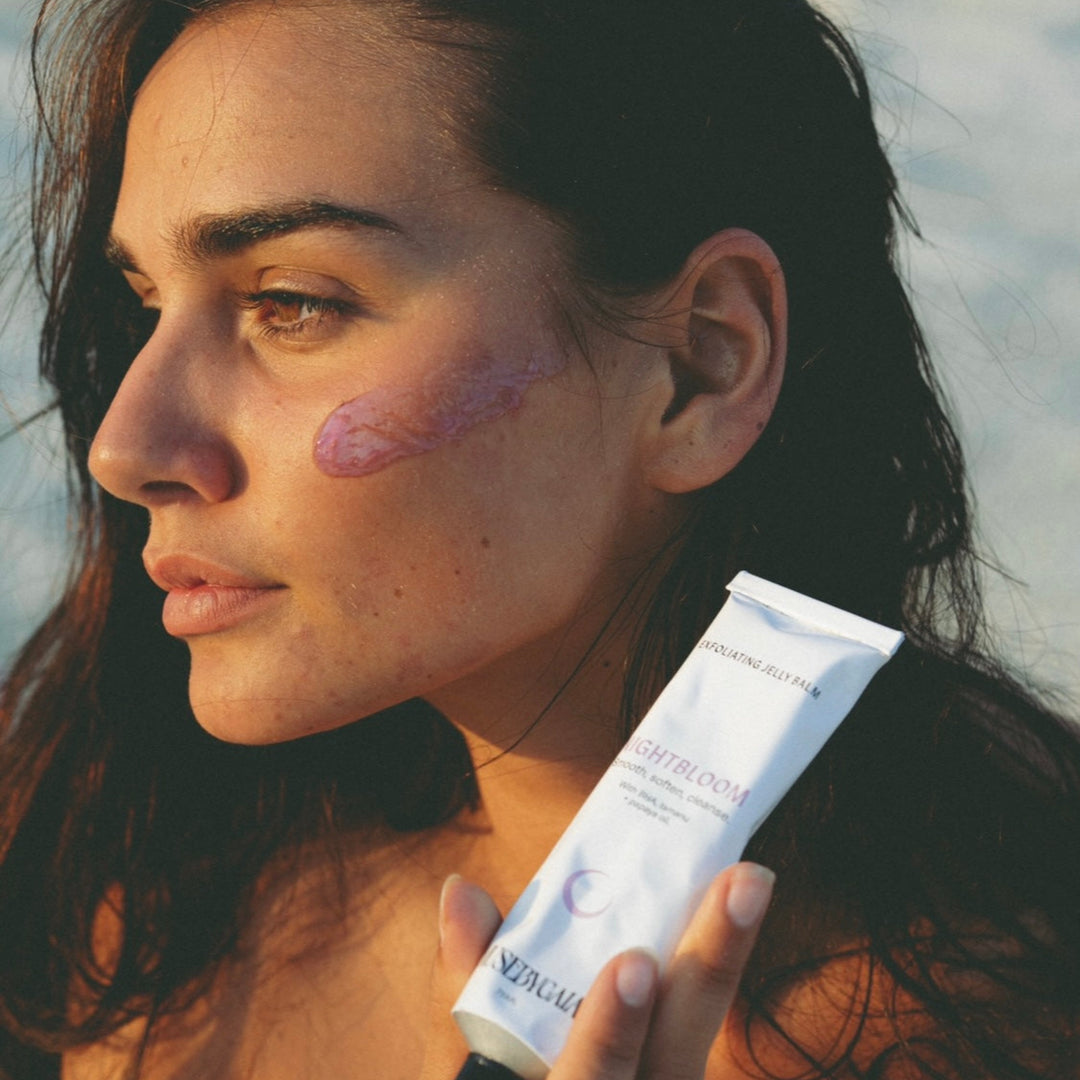 Profile view of a woman with dark hair at sunset, holding a tube of Muse by Gaia Nightbloom Exfoliating Jelly Balm and applying the pink gel on her cheek, emphasizing the product's real-time application and skin benefits.