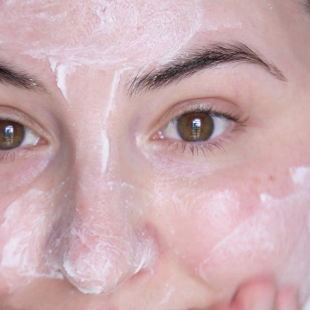 Close-up of a person's face with a creamy exfoliating mask applied unevenly across the skin, focusing on the area around the eyes. The individual has hazel eyes and a relaxed expression, with the mask highlighting the natural texture and tone of the skin.