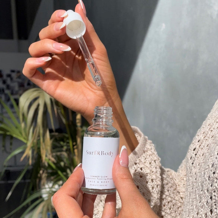 Woman holding a dropper and bottle of Surf & Body Summer Glow Hyaluronic Tanning Drops, preparing to apply the lightweight tanning serum for face and body.